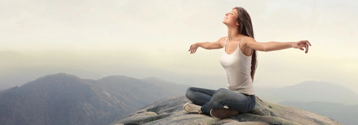 Person seated cross-legged on a rocky surface with arms extended and mountains in the background.