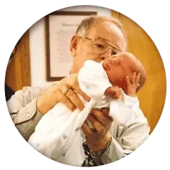 Close-up of a person holding an infant wrapped in a white blanket indoors.